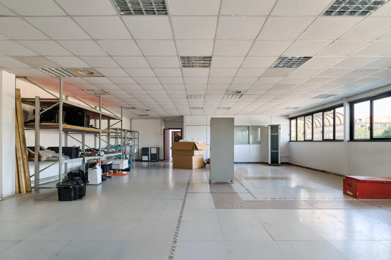 Ufficio spazioso, per lo più vuoto, con scaffali, scatole, un armadio, ampie finestre e oggetti sparsi sul pavimento.