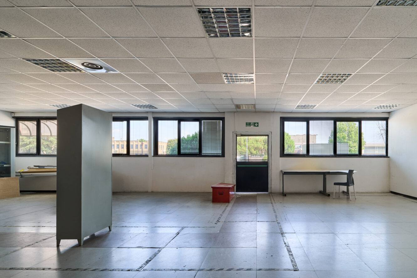 Un ufficio spazioso, quasi completamente vuoto, con il soffitto piastrellato, ampie finestre, una scrivania e una sedia, una scatola rossa e un armadio alto. La luce naturale inonda la stanza.