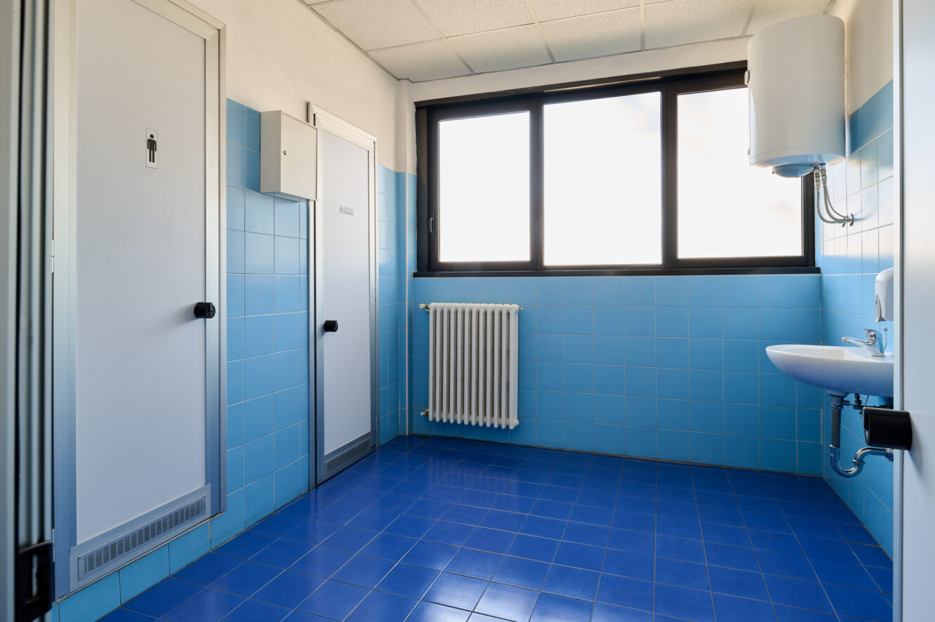 Bagno pubblico con pavimento e pareti piastrellati di blu, lavandino bianco, termosifone, scaldabagno, due porte che danno accesso ai box e un'ampia finestra che lascia entrare la luce naturale.