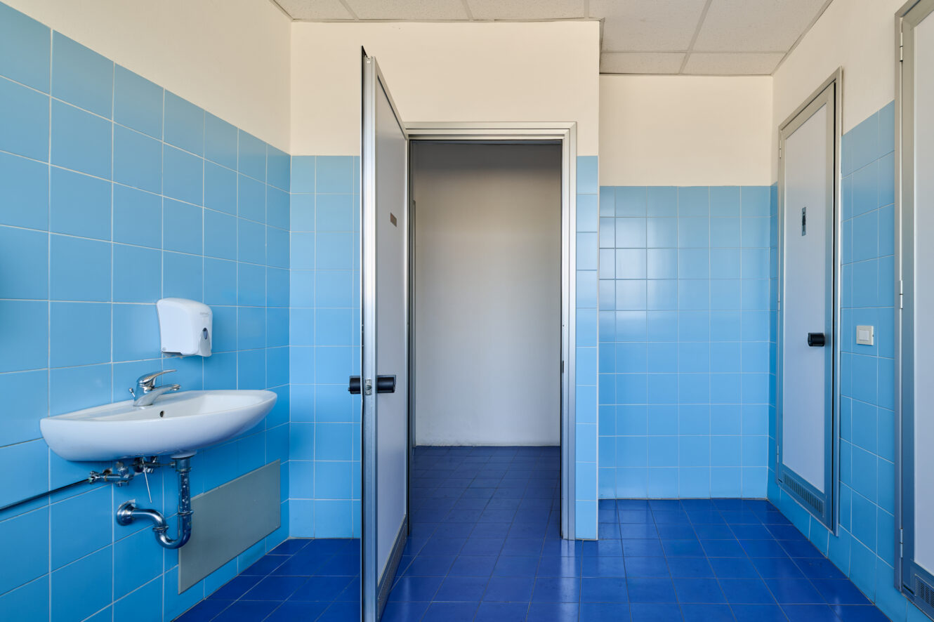 Un bagno pubblico con pareti e pavimento piastrellati di blu, un lavandino bianco con un dispenser di sapone e due porte di cabine, una aperta e una chiusa.