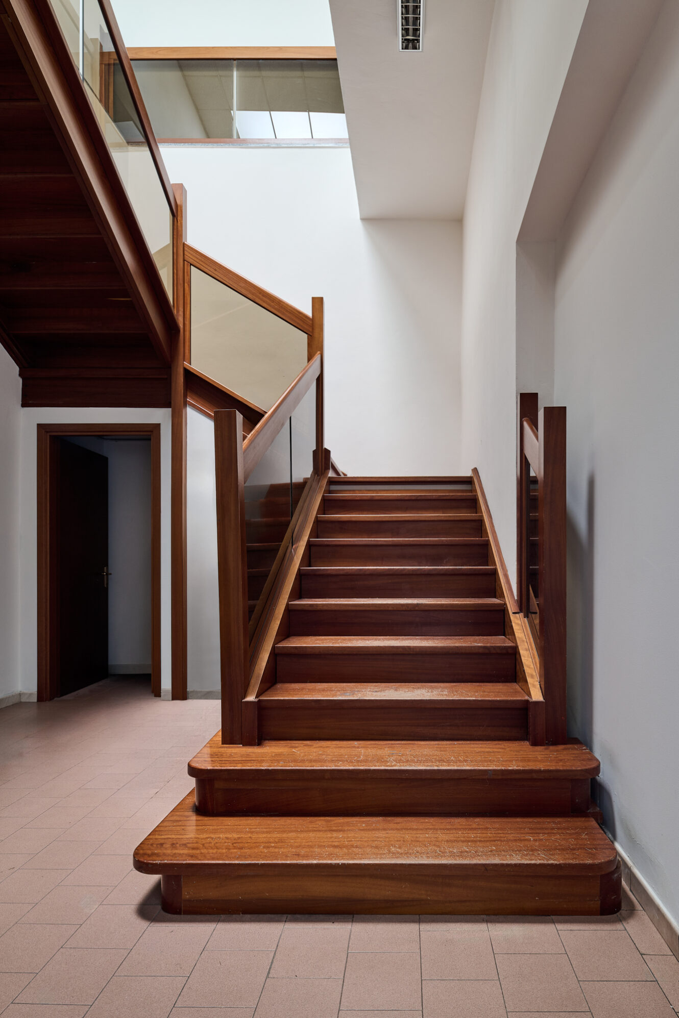 Una scala in legno con ringhiera a pannelli di vetro conduce a un pianerottolo in un ambiente moderno caratterizzato da pareti bianche e pavimento piastrellato. Alla base della scala si intravede una porta.