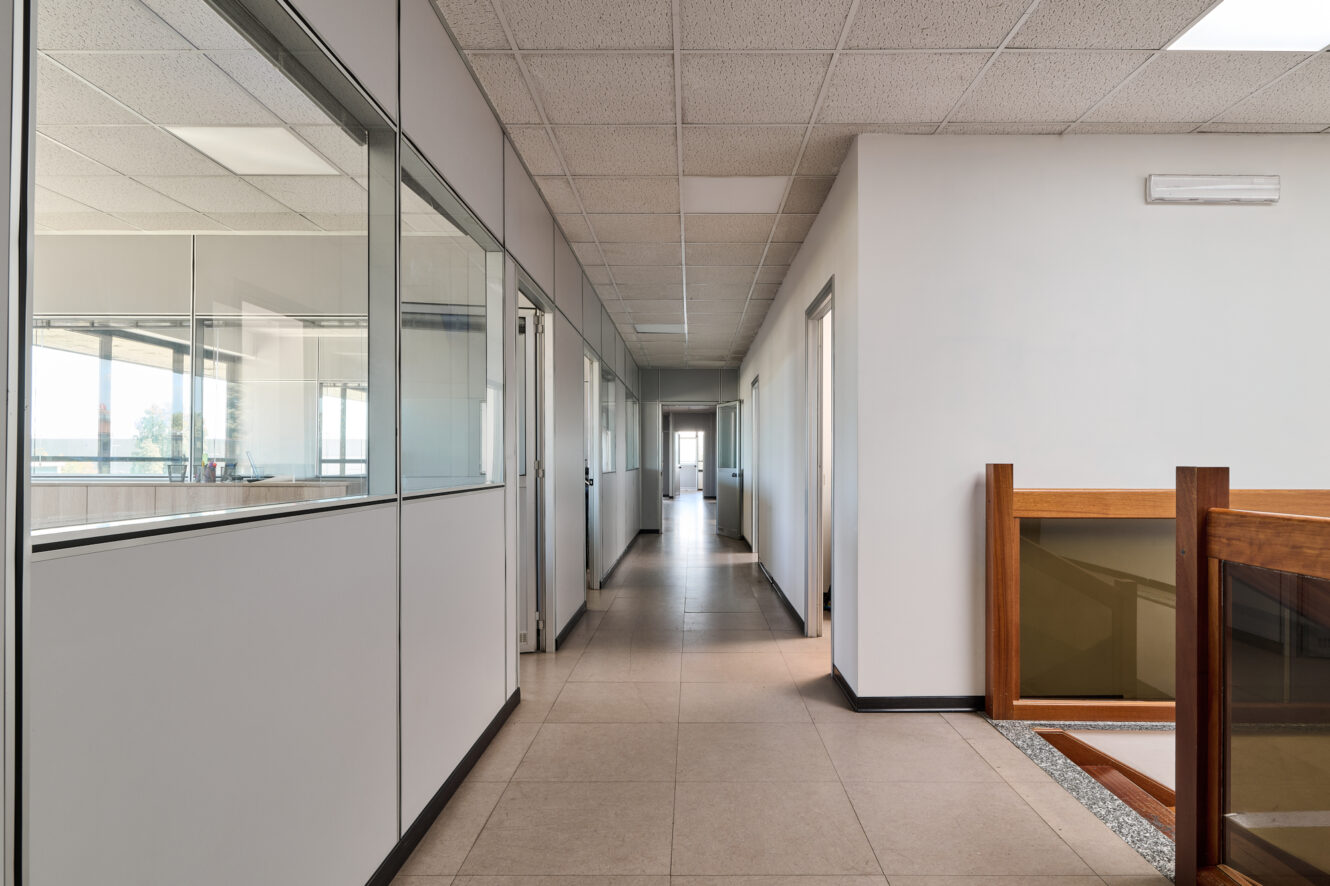 Un corridoio dell'ufficio pulito e vuoto, con stanze dalle pareti di vetro sulla sinistra, pareti bianche, pavimenti piastrellati e una ringhiera in legno della scala sulla destra.