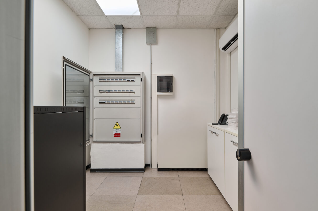 Piccola sala server con quadri elettrici, un rack per server nero, armadi bianchi, un telefono, pavimento piastrellato e pareti bianche illuminate da luci fluorescenti.