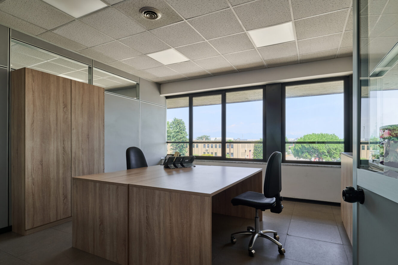 Ufficio moderno dotato di una spaziosa scrivania in legno, due sedie da ufficio, un telefono, armadietti e ampie finestre che offrono una vista su alberi ed edifici sotto un cielo sereno.