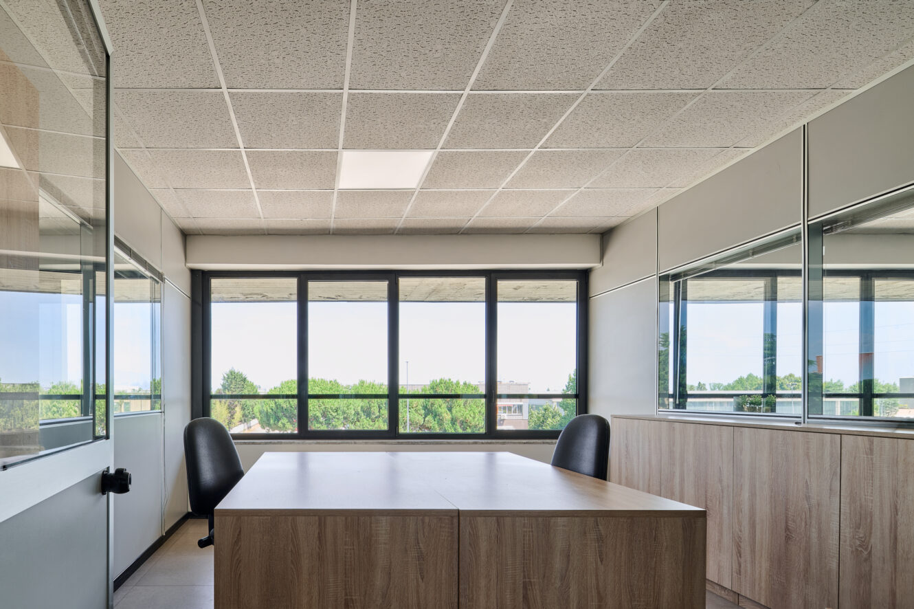 Ufficio luminoso e moderno con due sedie, una scrivania in legno e ampie finestre che si affacciano sugli alberi all'esterno. Lo spazio è caratterizzato da pareti dai toni neutri, pannelli per il soffitto e pareti divisorie in vetro.