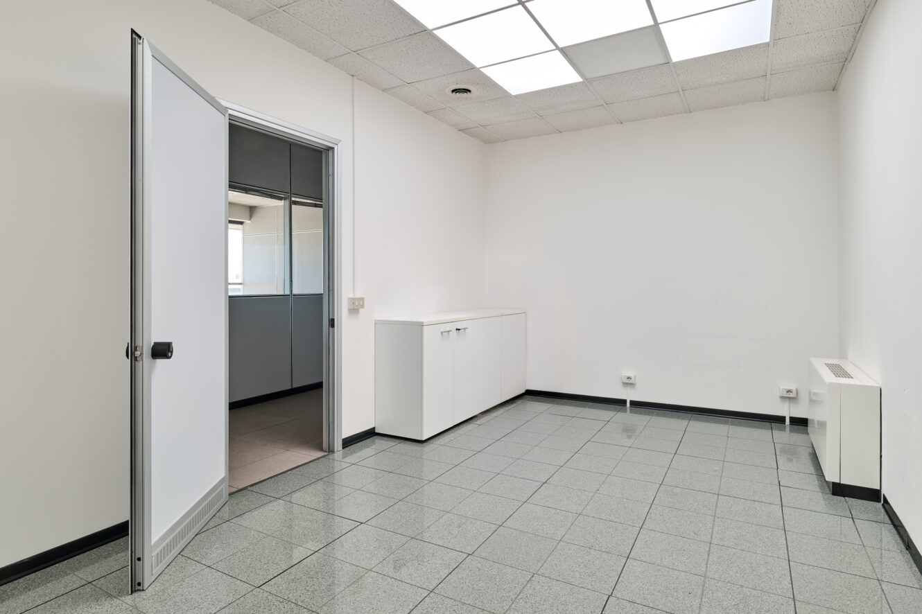 Ufficio vuoto con pavimento piastrellato, pareti bianche, luci fluorescenti a soffitto, una porta aperta, un armadio bianco e prese elettriche a vista.