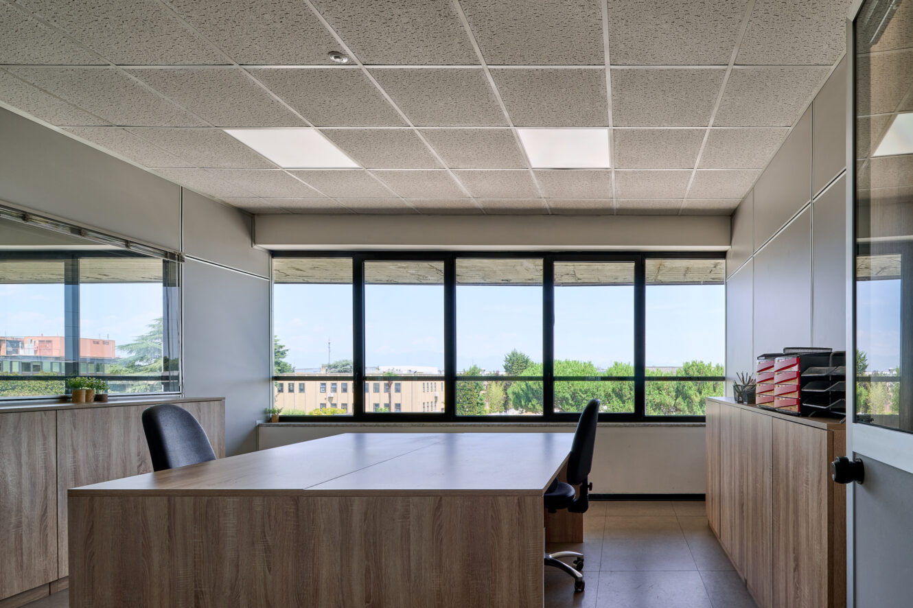 Ufficio moderno dotato di una scrivania in legno, due sedie, armadietti e ampie finestre con vista sugli alberi e sugli edifici. Lo spazio è inondato di luce naturale.