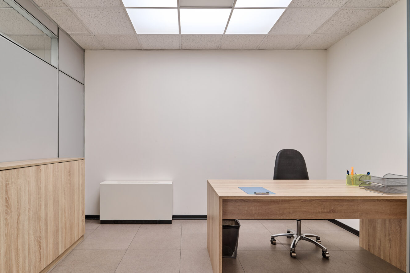 Ufficio in stile minimalista con scrivania in legno, sedia nera, armadio chiuso e materiale da scrittura disposto ordinatamente sulla scrivania; pareti bianche e pavimento piastrellato.