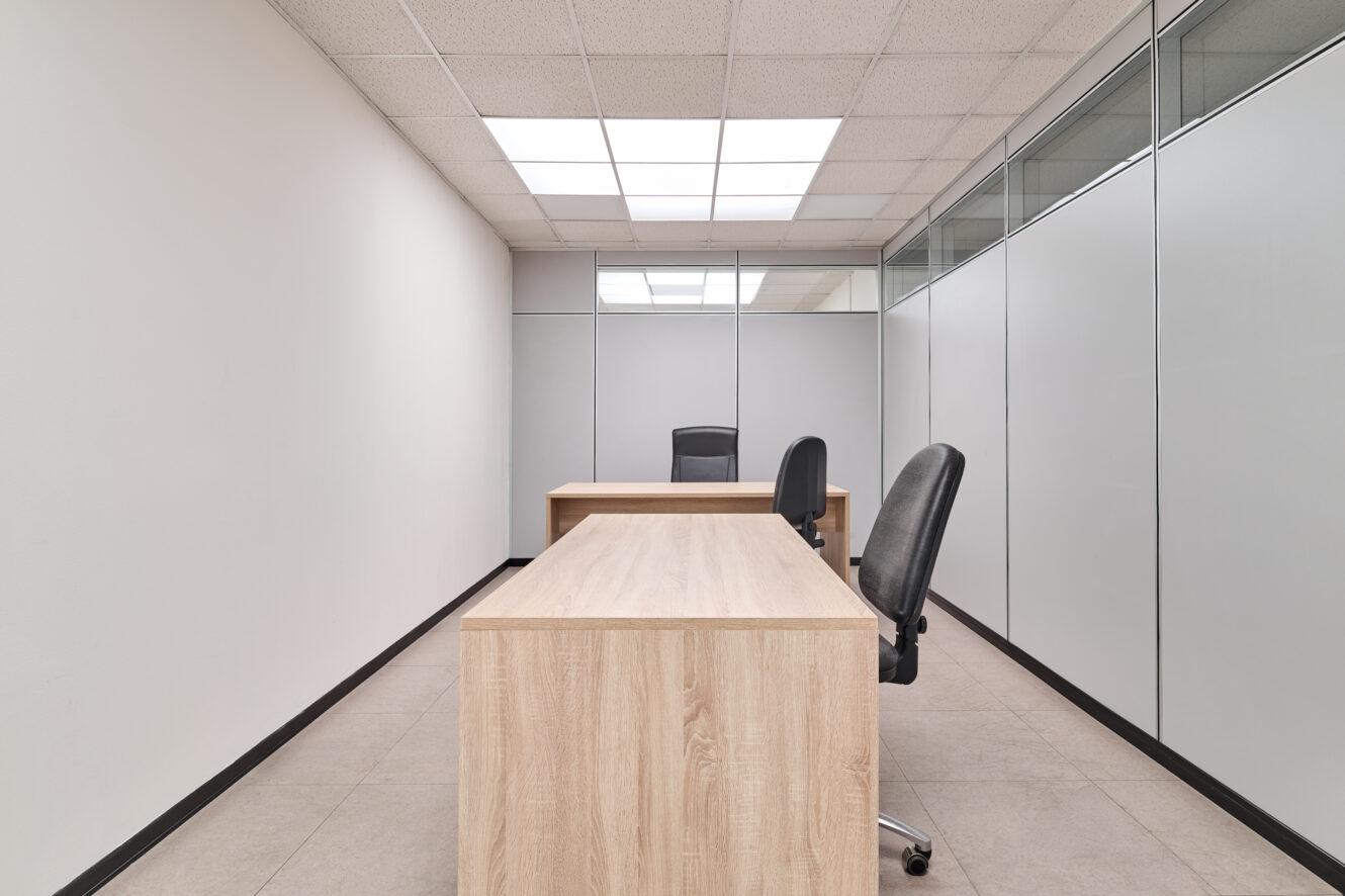 Ufficio in stile minimalista con due scrivanie in legno, due sedie nere, pavimento piastrellato, pareti bianche e divisori in vetro sotto le luci fluorescenti a soffitto.