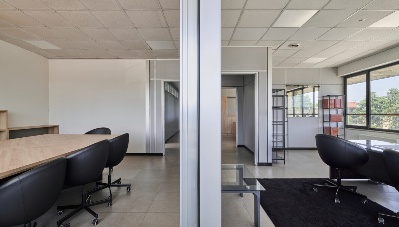 Moderno spazio ufficio suddiviso da una parete di vetro, con tavoli da riunione, sedie nere, finestre con vista sull’esterno e arredamento minimalista.
