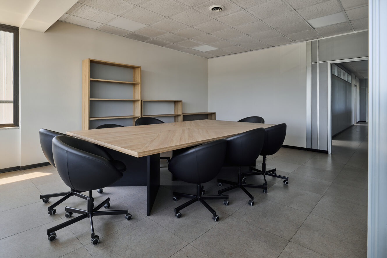 Sala riunioni con un tavolo in legno, otto sedie nere su rotelle, scaffali vuoti, pavimento piastrellato e ampie finestre che lasciano entrare la luce naturale.