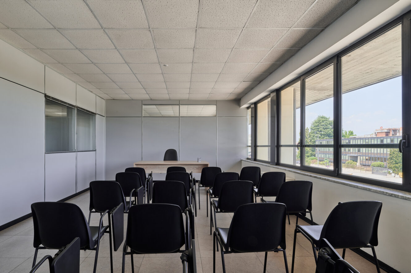 Aula vuota con file di sedie nere rivolte verso una cattedra e una sedia in fondo alla classe, grandi finestre sulla destra e pareti bianche con armadietti sulla sinistra.