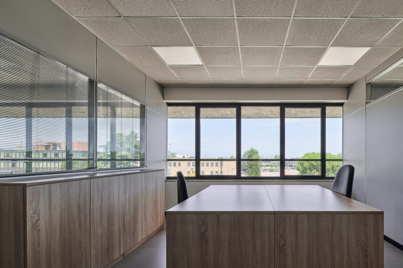 Ufficio moderno con due sedie nere, una scrivania in legno, ampie finestre e armadietti. Lo spazio è vuoto e si affaccia sugli edifici e sugli alberi all’esterno.