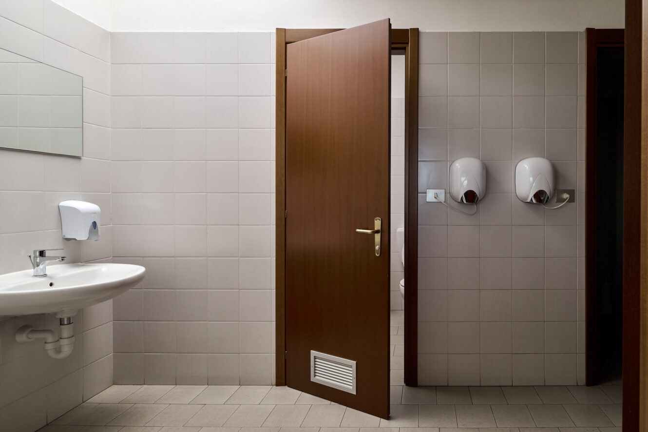 Un bagno pubblico dotato di un lavandino bianco, un dispenser di sapone, uno specchio rettangolare, due asciugamani elettrici a parete e una porta in legno parzialmente aperta.