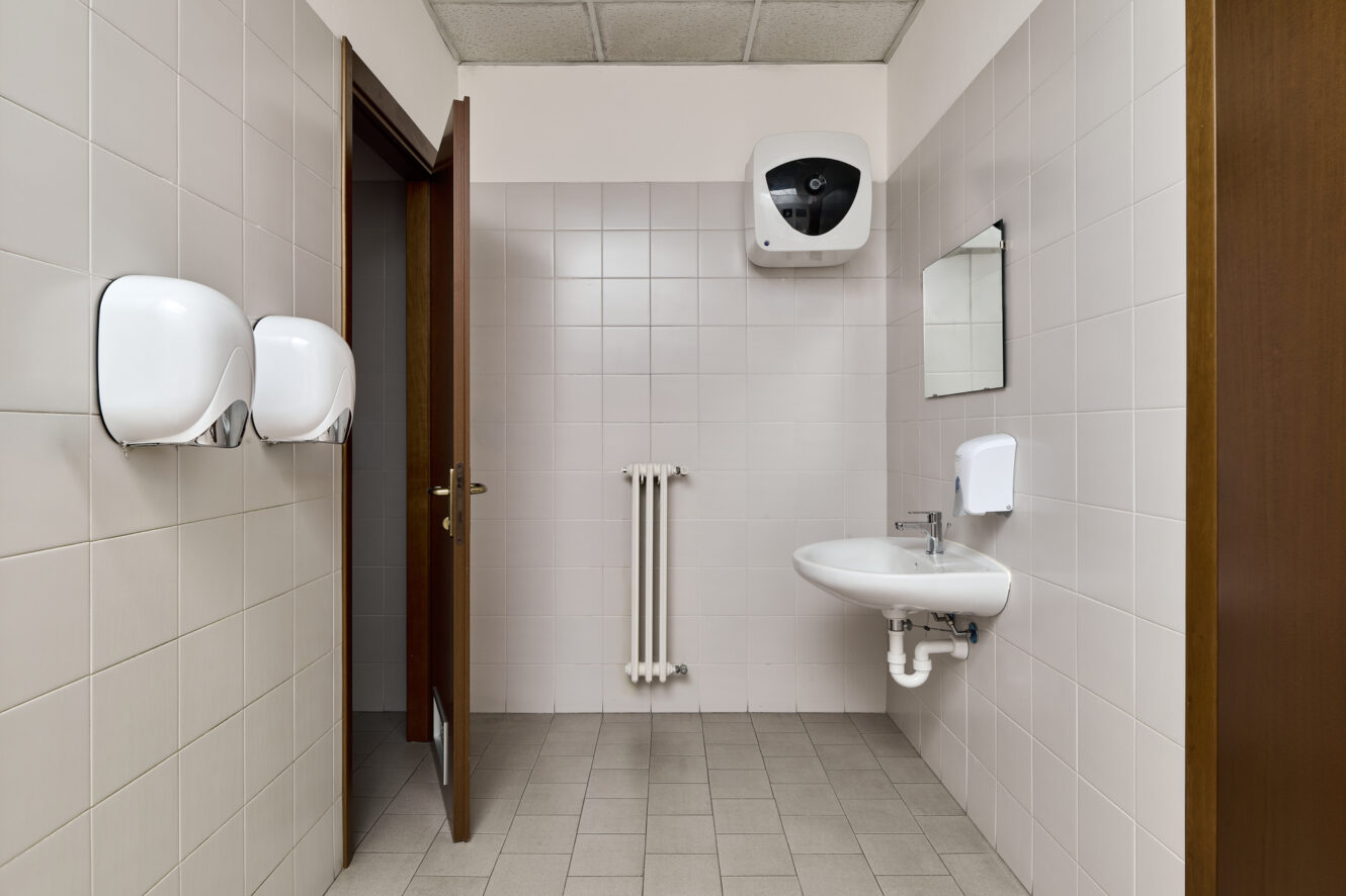 Bagno pubblico con pareti piastrellate, porta aperta, lavandino a muro, specchio, dispenser di sapone, due dispenser di asciugamani di carta, termosifone e un asciugamani elettrico sopra lo specchio.