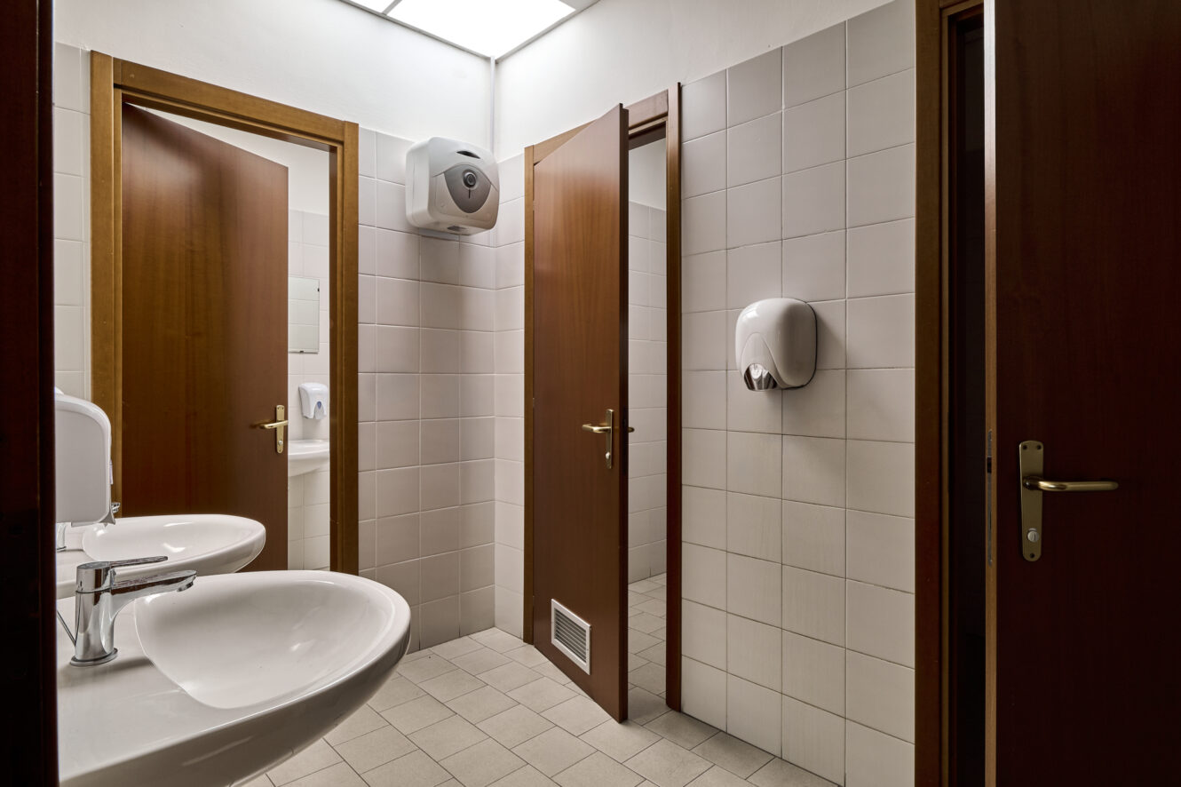 Bagno pubblico con pareti piastrellate di bianco, tre porte in legno, due lavandini, un dispenser di sapone e un dispenser di asciugamani di carta ben visibili.
