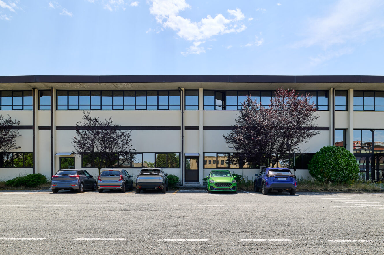 Quattro auto, tra cui un’auto sportiva verde, sono parcheggiate davanti a un edificio di due piani adibito a uffici, con ampie vetrate e alcuni alberi.
