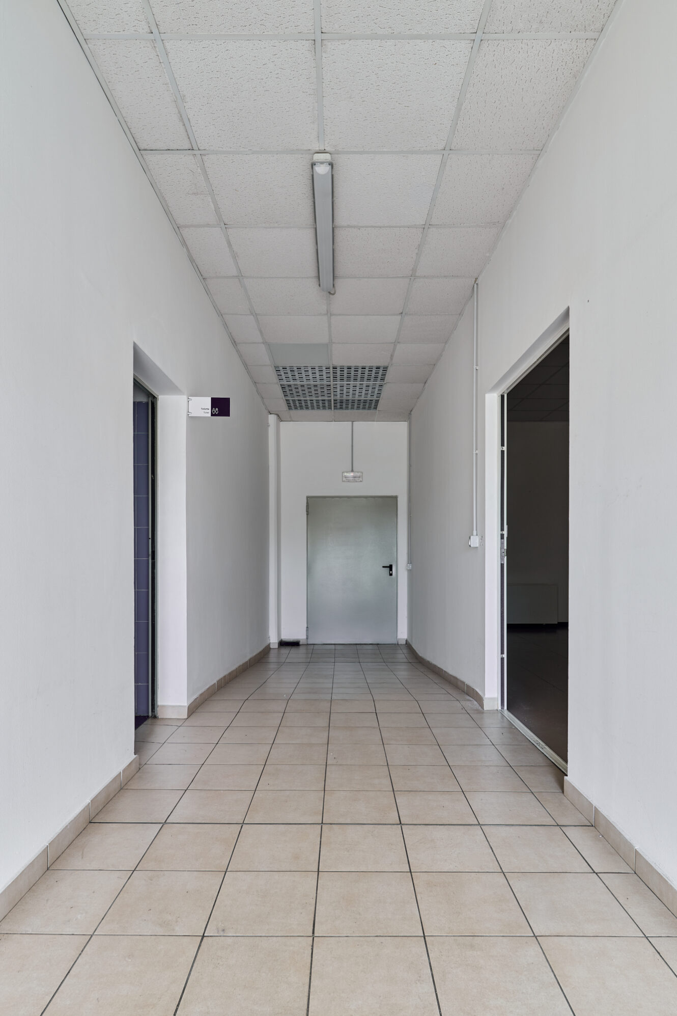 Un corridoio pulito e vuoto, con pavimento piastrellato, pareti bianche, luci fluorescenti a soffitto e una porta grigia chiusa in fondo. Su entrambi i lati si vedono due porte aperte.