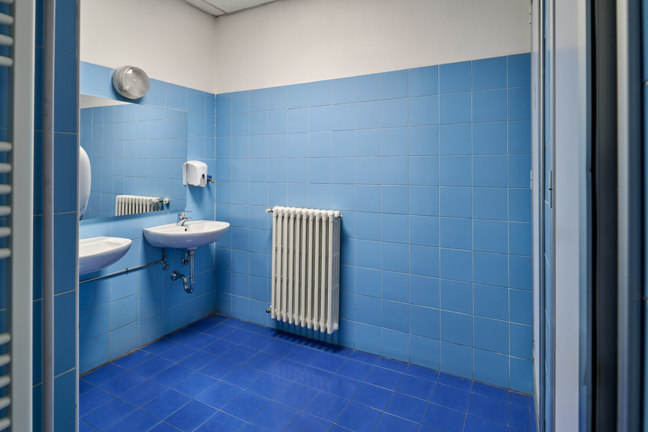 Un bagno con piastrelle blu, un lavandino a muro, uno specchio, un termosifone, un dispenser per il sapone e una lampada da parete rotonda.