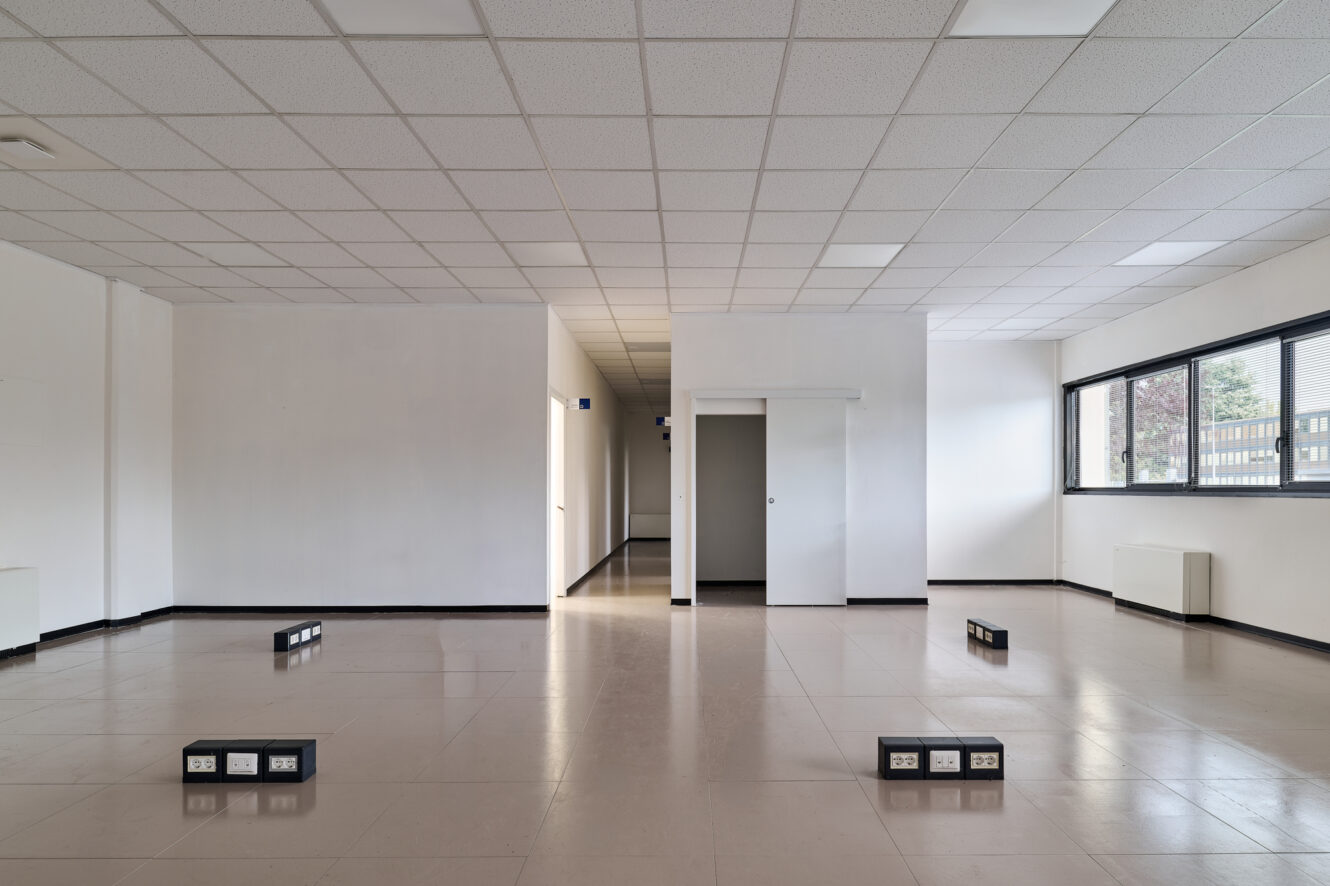Spazio ufficio vuoto con pareti bianche, controsoffitto, pavimento piastrellato, diverse prese elettriche a pavimento e ampie finestre sulla destra che lasciano entrare la luce naturale.