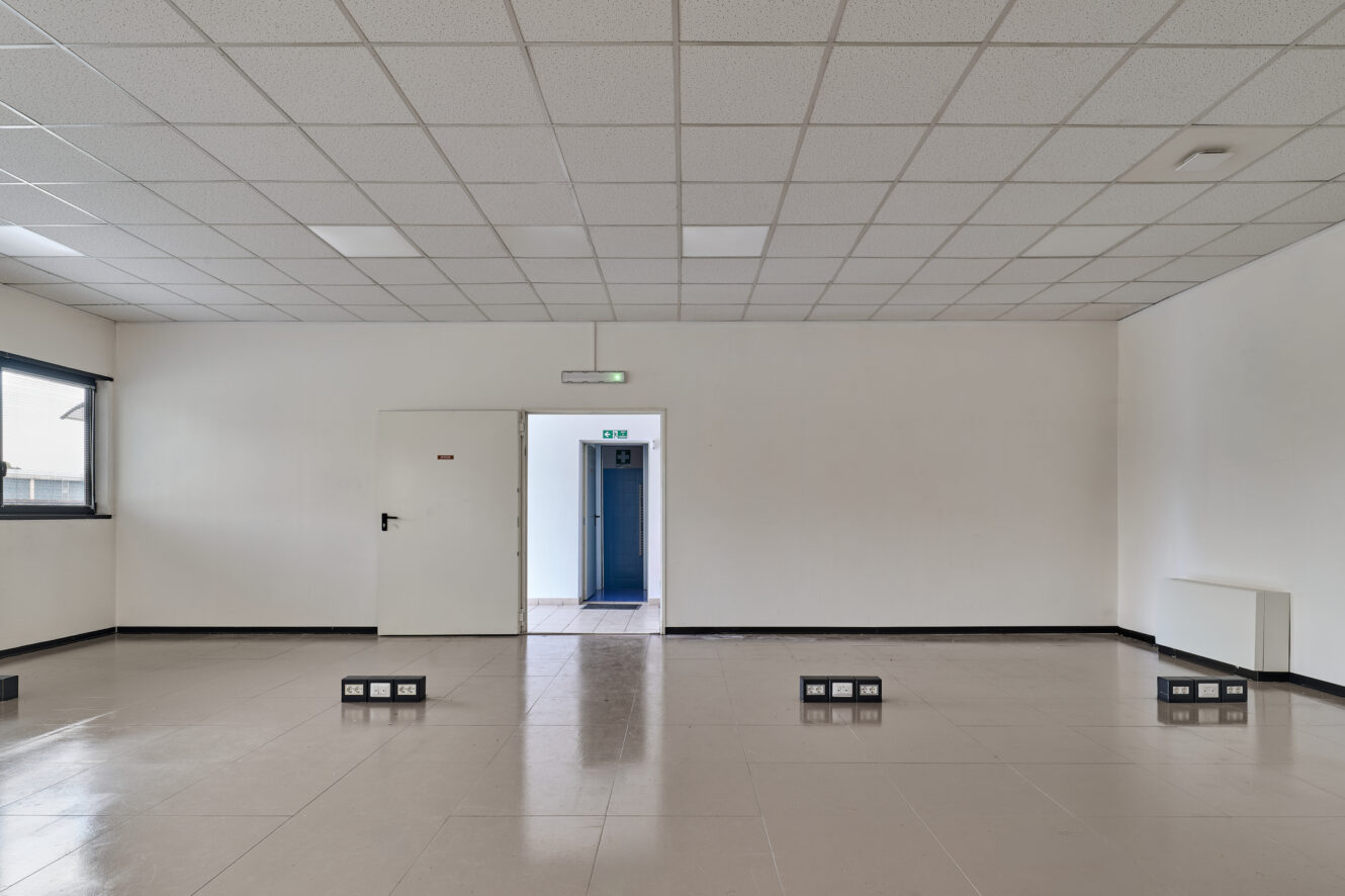 Stanza vuota con pareti bianche, soffitto piastrellato, prese a pavimento, una finestra chiusa, una porta bianca e una porta aperta che conduce a un corridoio blu.