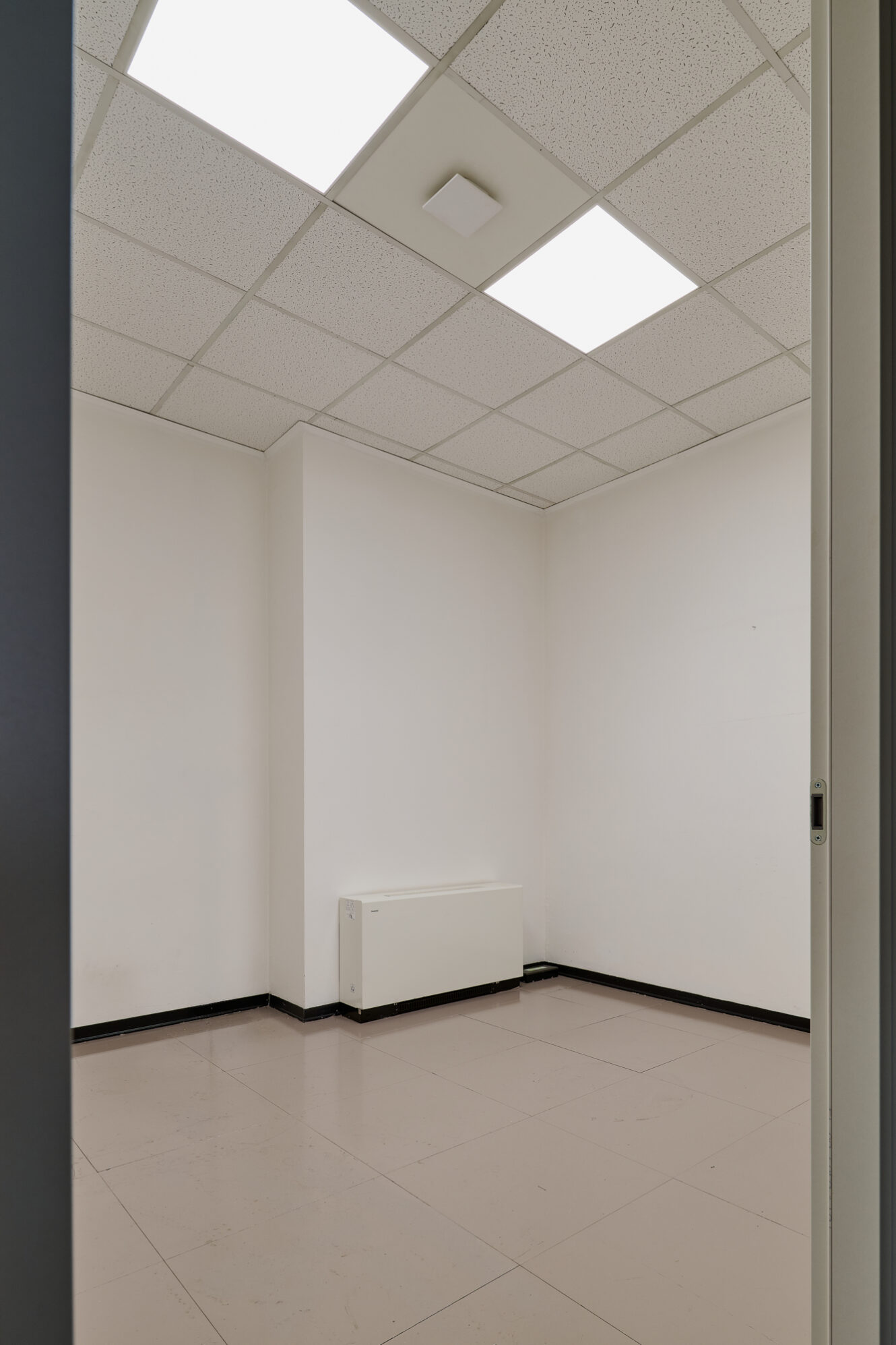Piccolo ufficio vuoto con pareti bianche, pavimento piastrellato, controsoffitto con luci fluorescenti e un termosifone a parete.