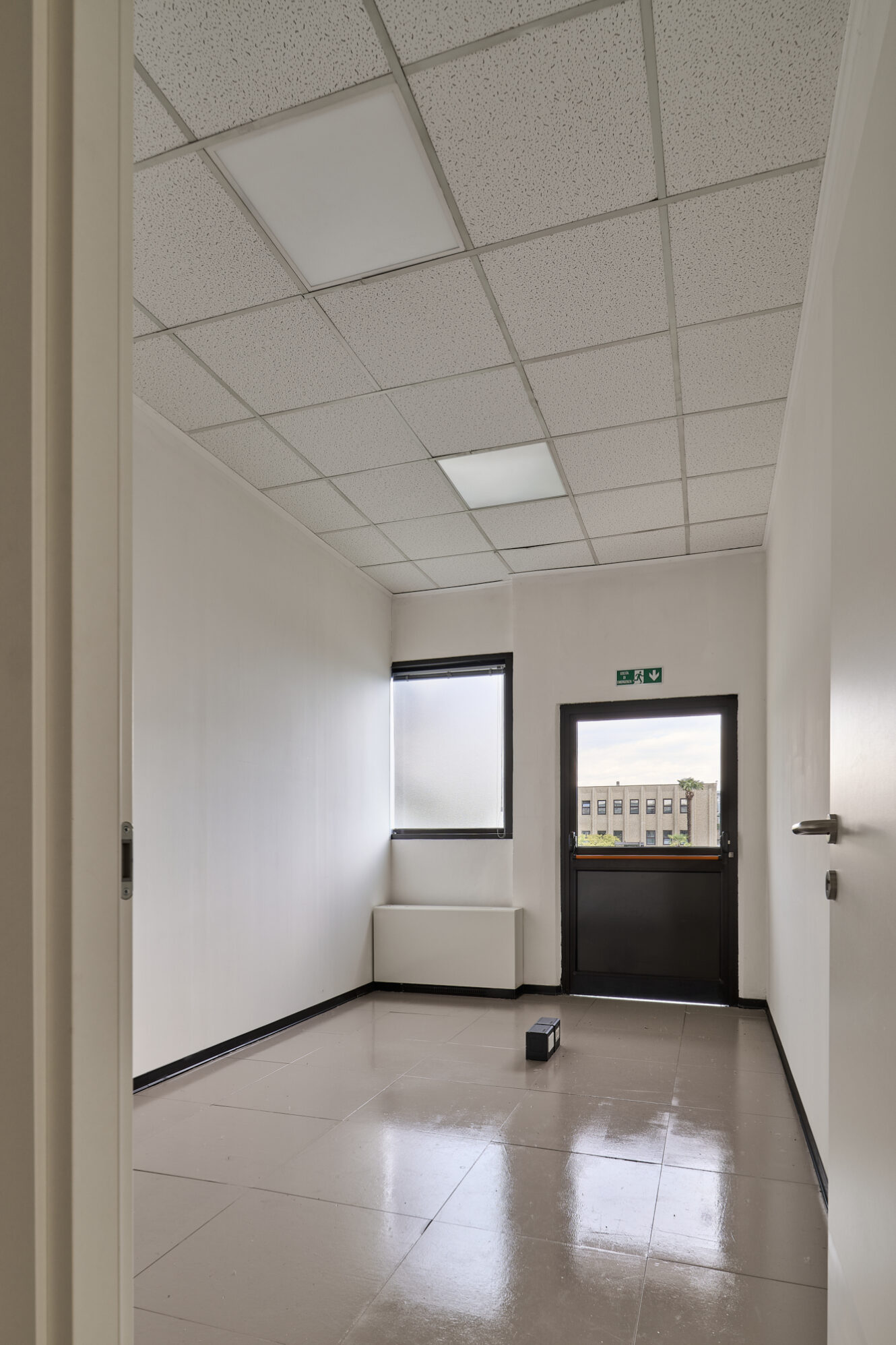Stanza da ufficio vuota e non arredata, con soffitto piastrellato, una finestra, una porta nera con un cartello di uscita e un'unica scatola nera sul pavimento lucido.