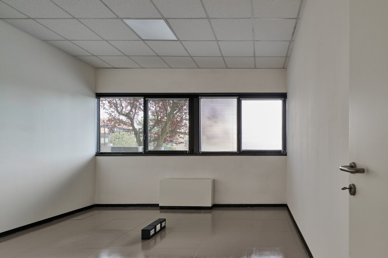 Stanza da ufficio vuota e non arredata, con pareti bianche, pavimento piastrellato, tre finestre e una lampada da soffitto rettangolare; un piccolo oggetto nero giace sul pavimento vicino al centro.