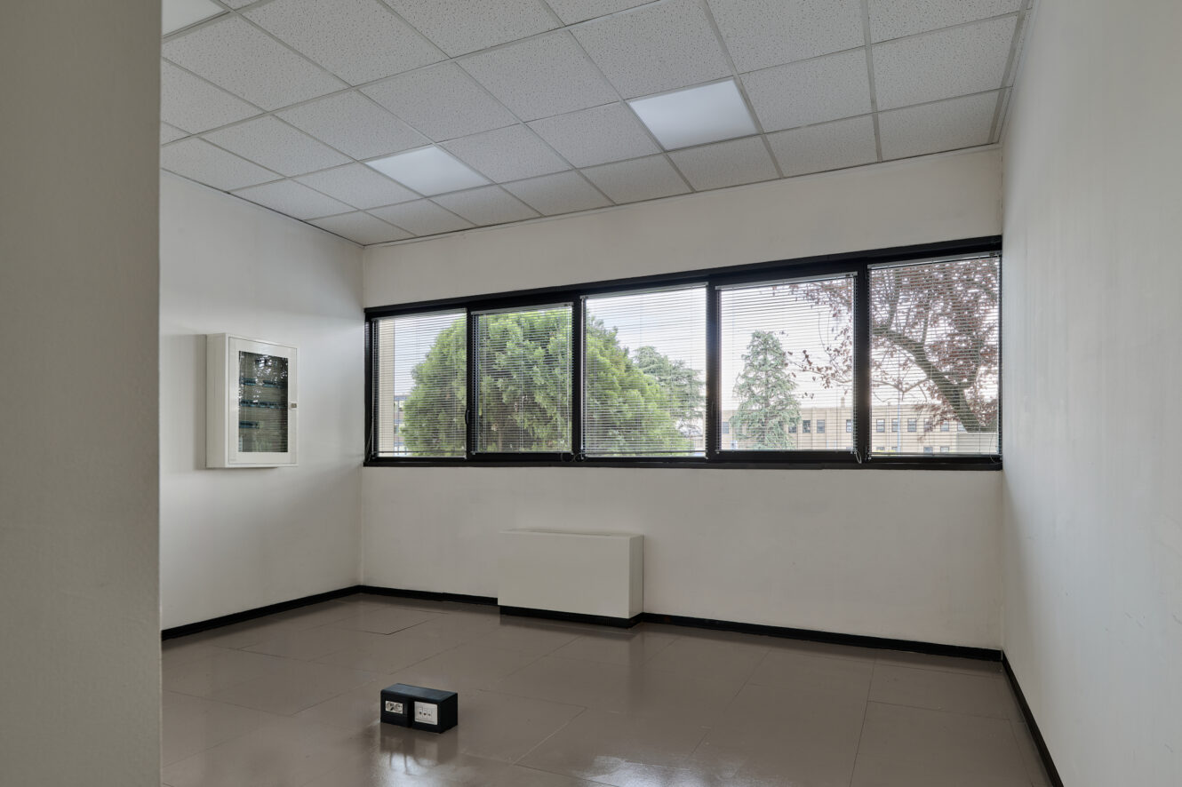 Ufficio vuoto con ampie finestre, una piccola scatola nera sul pavimento piastrellato, pareti bianche e un soffitto con luci fluorescenti. Fuori dalle finestre si vedono degli alberi.