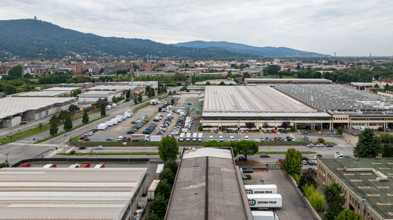 Vista aerea di una zona industriale con magazzini, veicoli parcheggiati e montagne sullo sfondo, sotto un cielo nuvoloso.