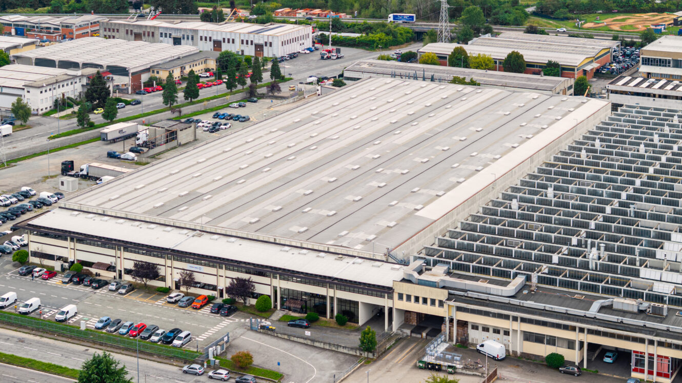 Vista aerea di un grande complesso di capannoni industriali con camion, auto parcheggiate ed edifici adiacenti, circondato da strade e aree verdi.