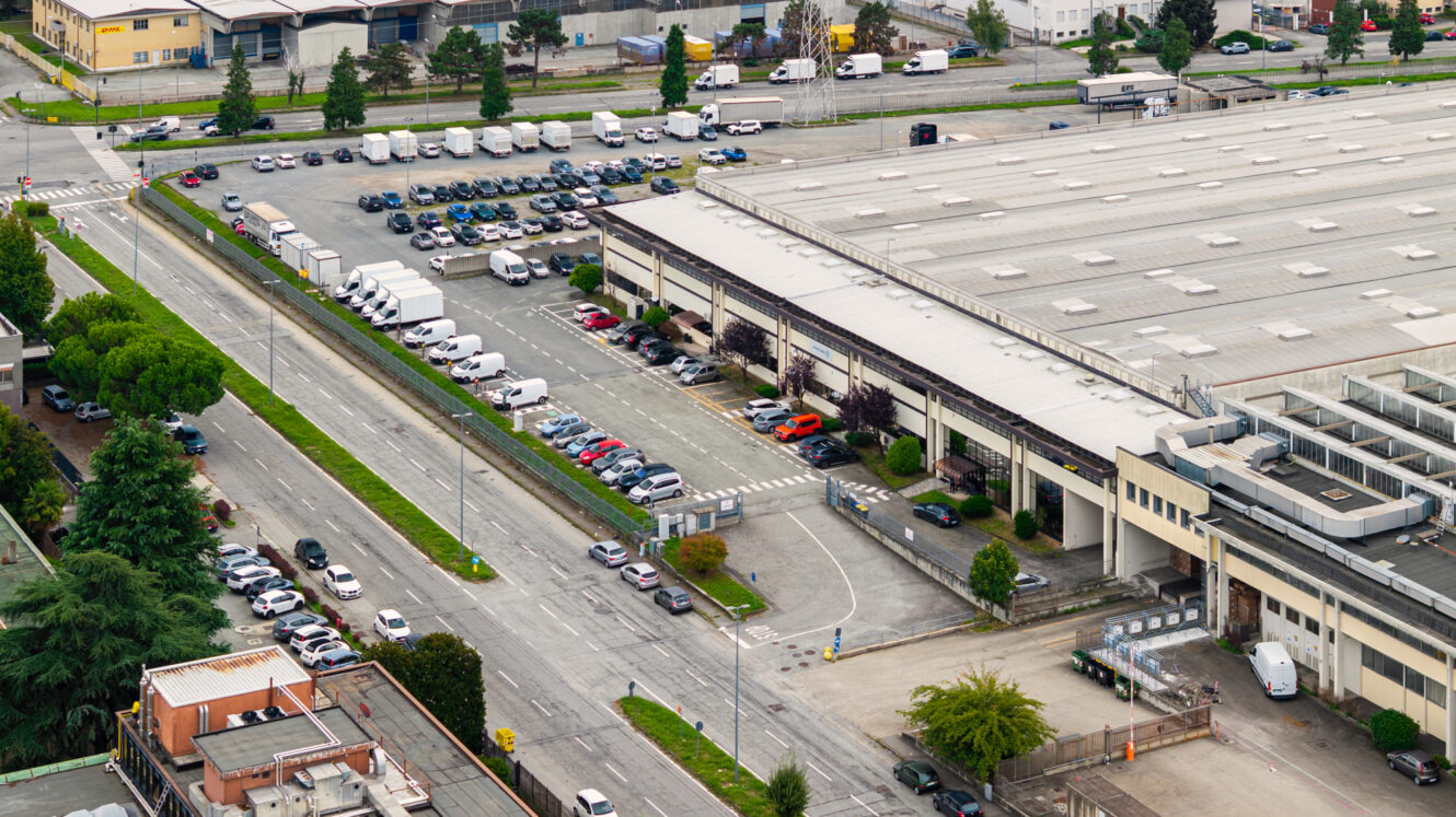 Vista aerea di un edificio industriale con un ampio parcheggio, diversi veicoli e le strade circostanti in un’area urbana.