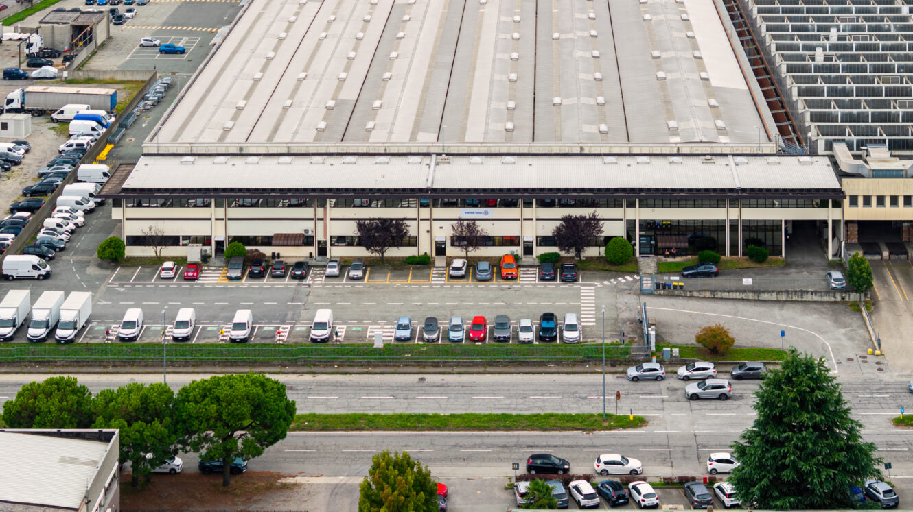 Vista aerea di un grande capannone industriale con auto e furgoni di consegna parcheggiati davanti, circondato da strade e vegetazione.