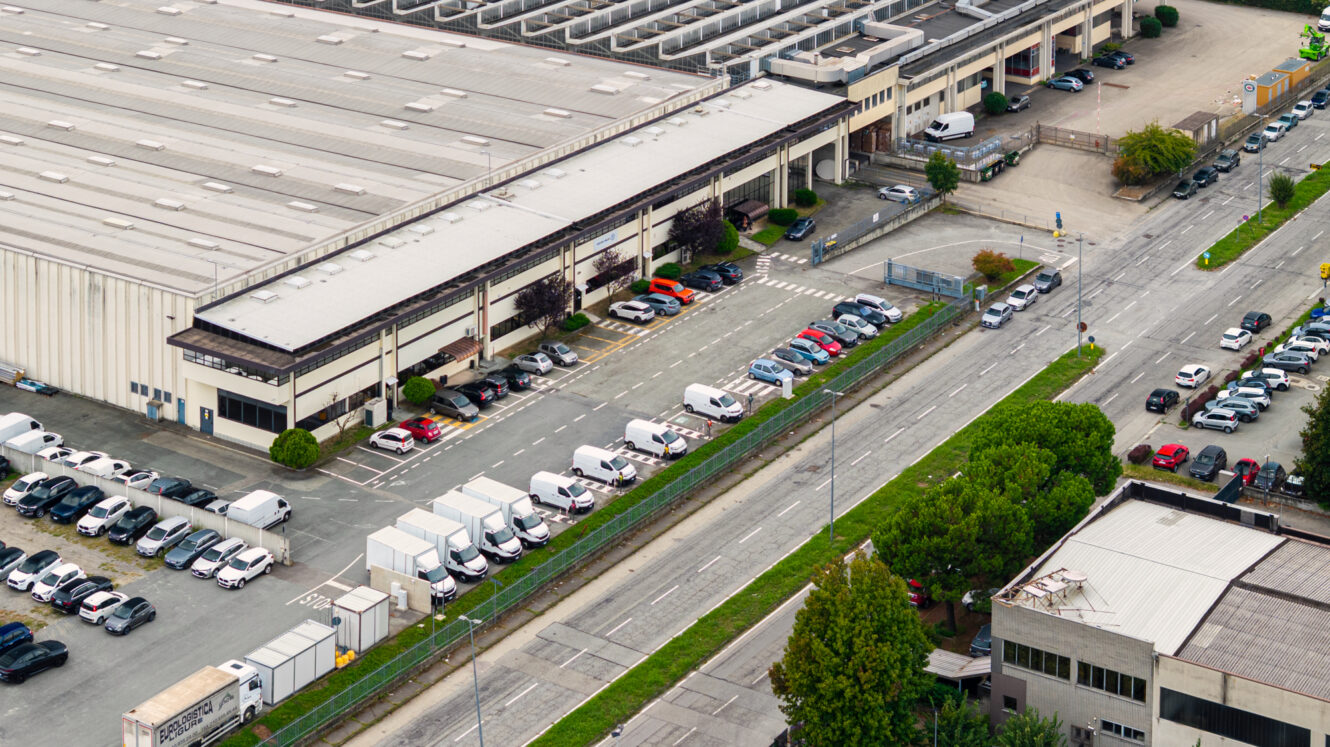 Vista aerea di un edificio industriale con un parcheggio pieno di auto e furgoni, circondato da strade, alberi e altre strutture.