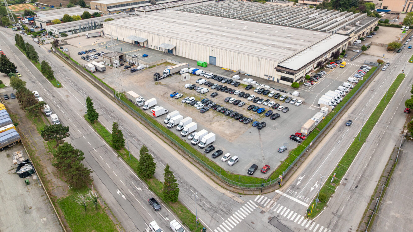 Vista aerea di un grande magazzino con un parcheggio adiacente in cui sono parcheggiate auto e furgoni, circondato da strade e dal verde.