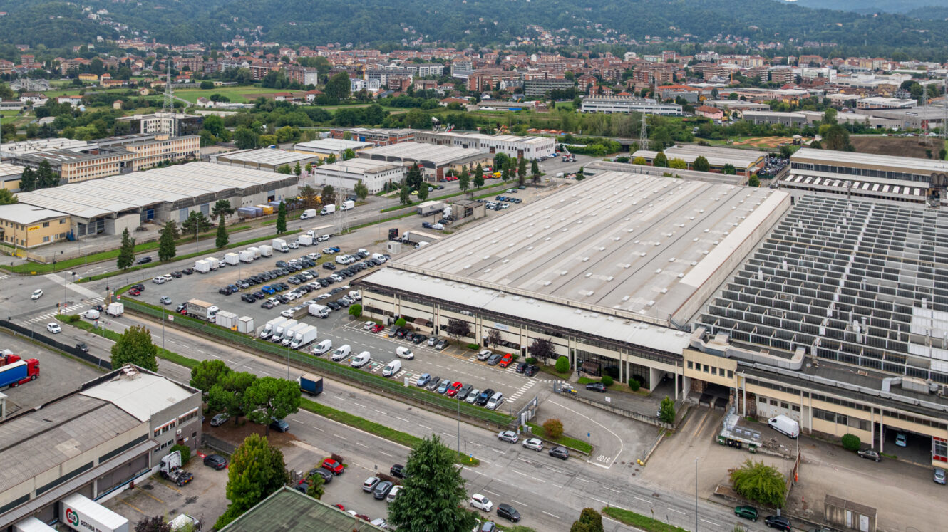 Vista aerea di una zona industriale con grandi magazzini, parcheggi, camion e strade circostanti fiancheggiate da alberi, con edifici residenziali sullo sfondo.