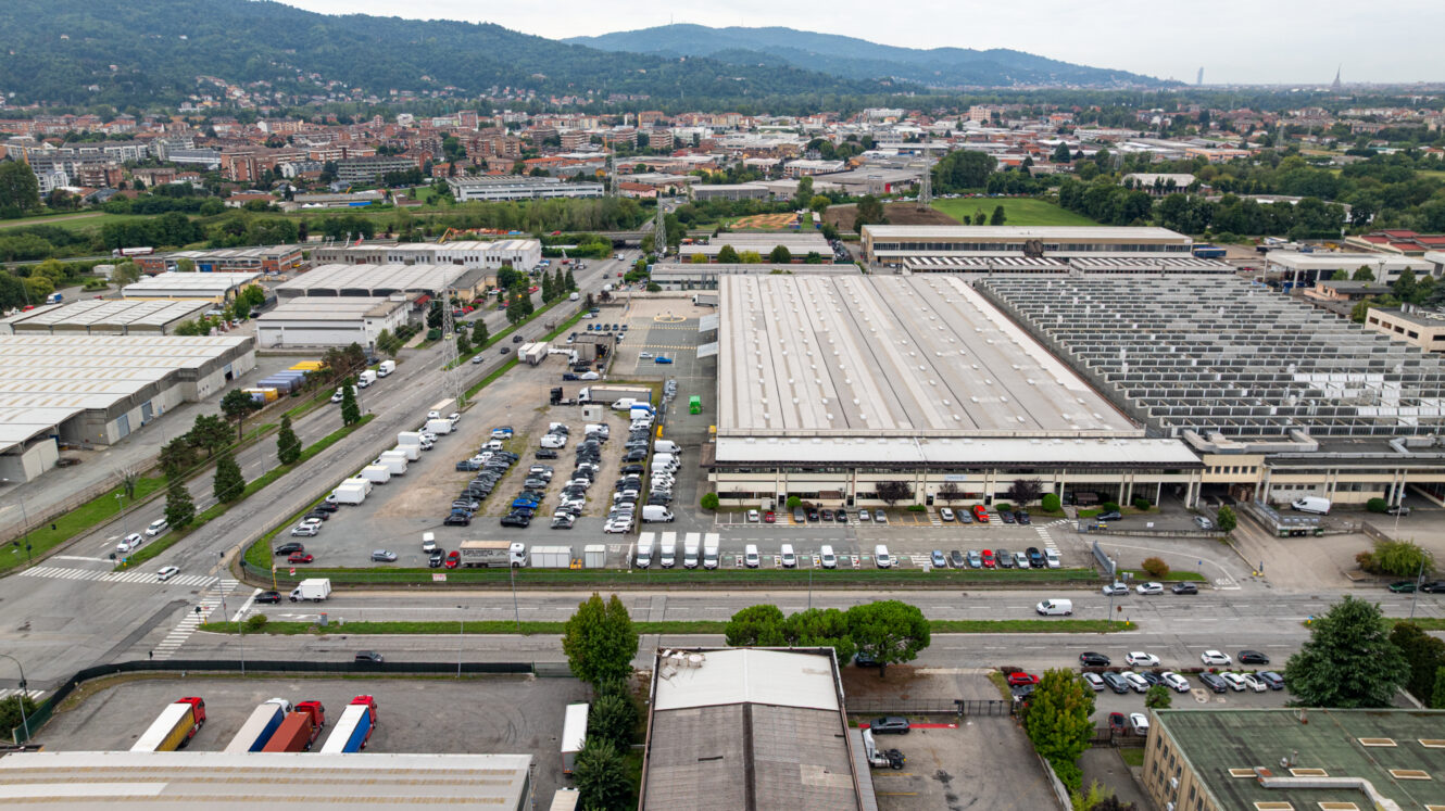 Vista aerea di un complesso industriale con magazzini, auto parcheggiate, camion e strade circostanti su cui si nota un certo viavai, il tutto sullo sfondo di colline e di un panorama urbano.