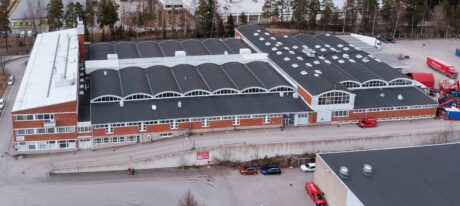 Ilmakuva suuresta teollisuus- tai varastorakennuksesta, jossa on musta katto, punatiiliset seinät, useita pysäköityjä ajoneuvoja ja puita taustalla.