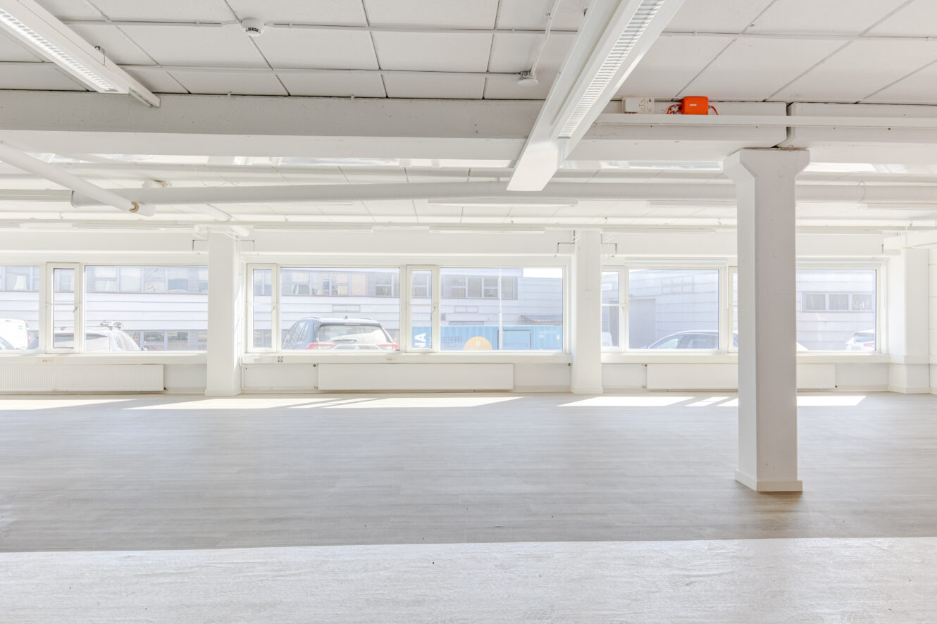 Valoisa, tyhjä liiketila, jossa on suuret ikkunat, valkoiset seinät, kattoputket ja betonilattiat. Ikkunoiden läpi näkyy useita pysäköityjä autoja.