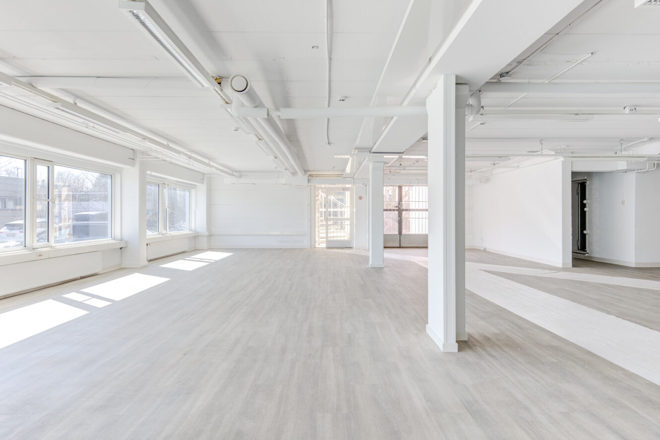 Tilava, tyhjä huone, jossa on suuret ikkunat, valkoiset seinät, vaalea puulattia ja kattoputket, täynnä luonnonvaloa.