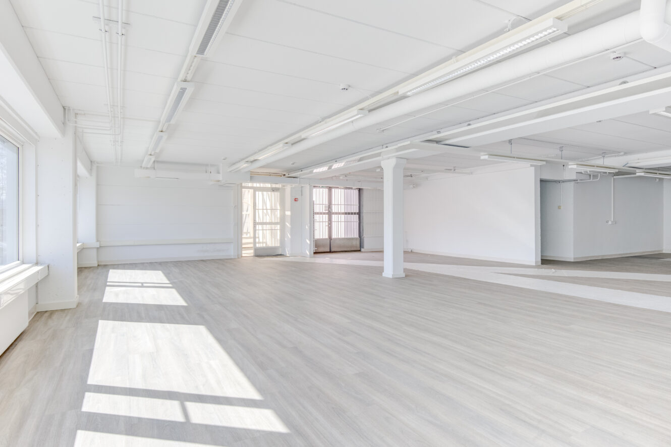 Tilava, tyhjä huone, jossa on suuret ikkunat, valkoiset seinät, pylväät ja vaalea puulattia, jota valaisee luonnonvalo.