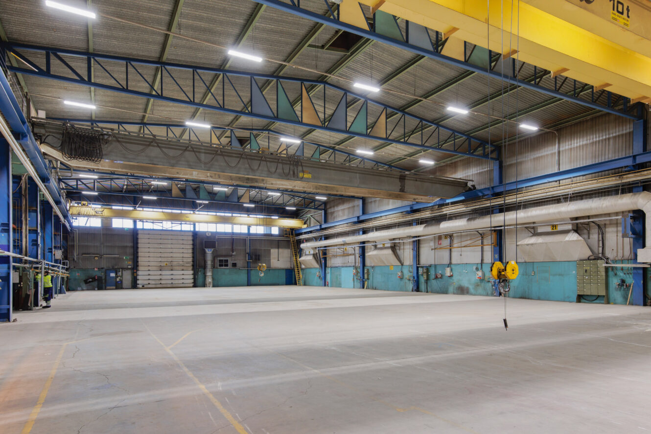 Suuri, tyhjä teollisuusvarasto, jossa on korkeat katot, nosturit, loisteputkivalot ja betonilattiat. Seinät on maalattu siniseksi ja valkoiseksi, ja tila on siisti ja hyvin valaistu.