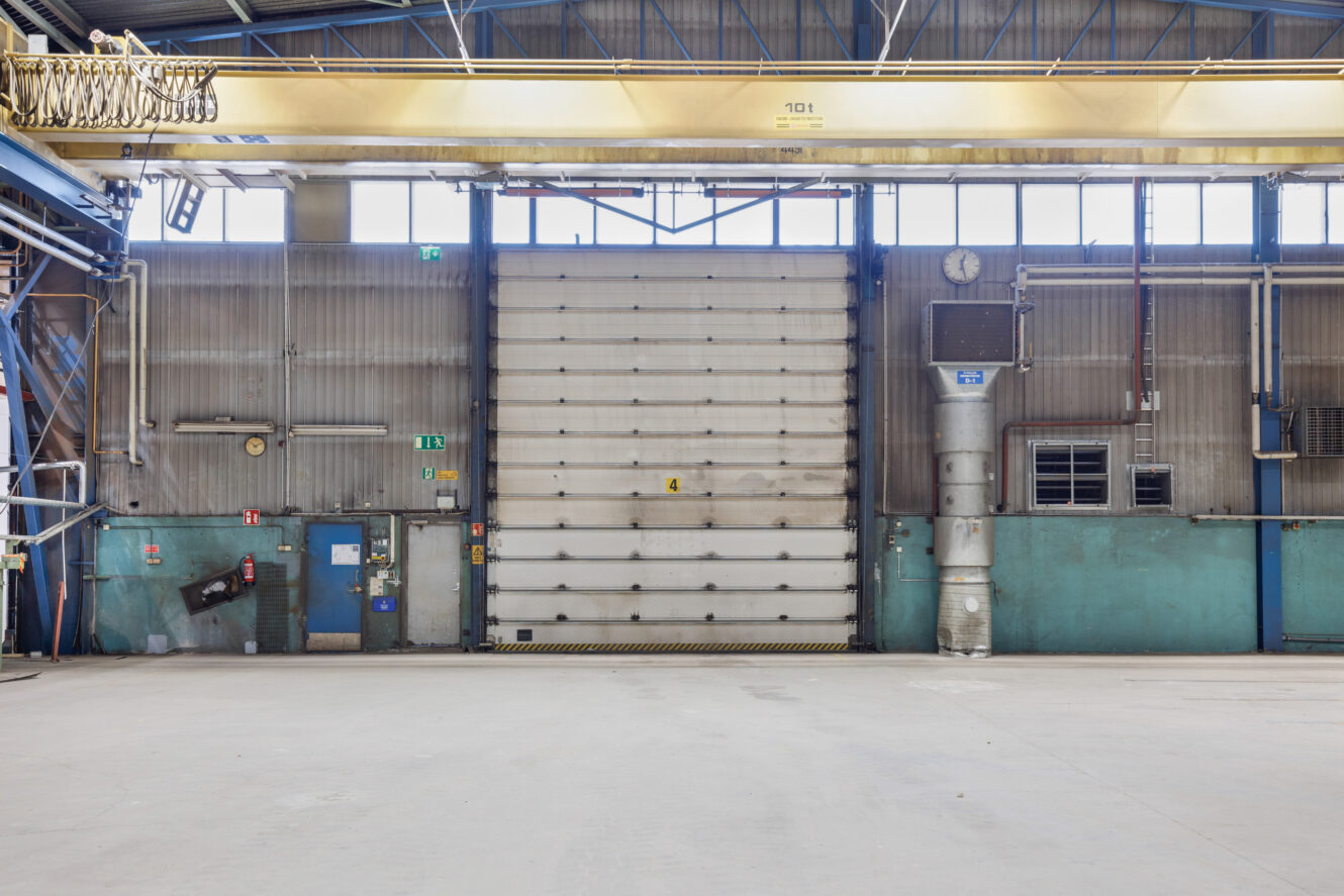 Tyhjän teollisuusvaraston sisätila, jossa on suuri suljettu rullaportti, nosturi, metalliset seinät ja betonilattia.