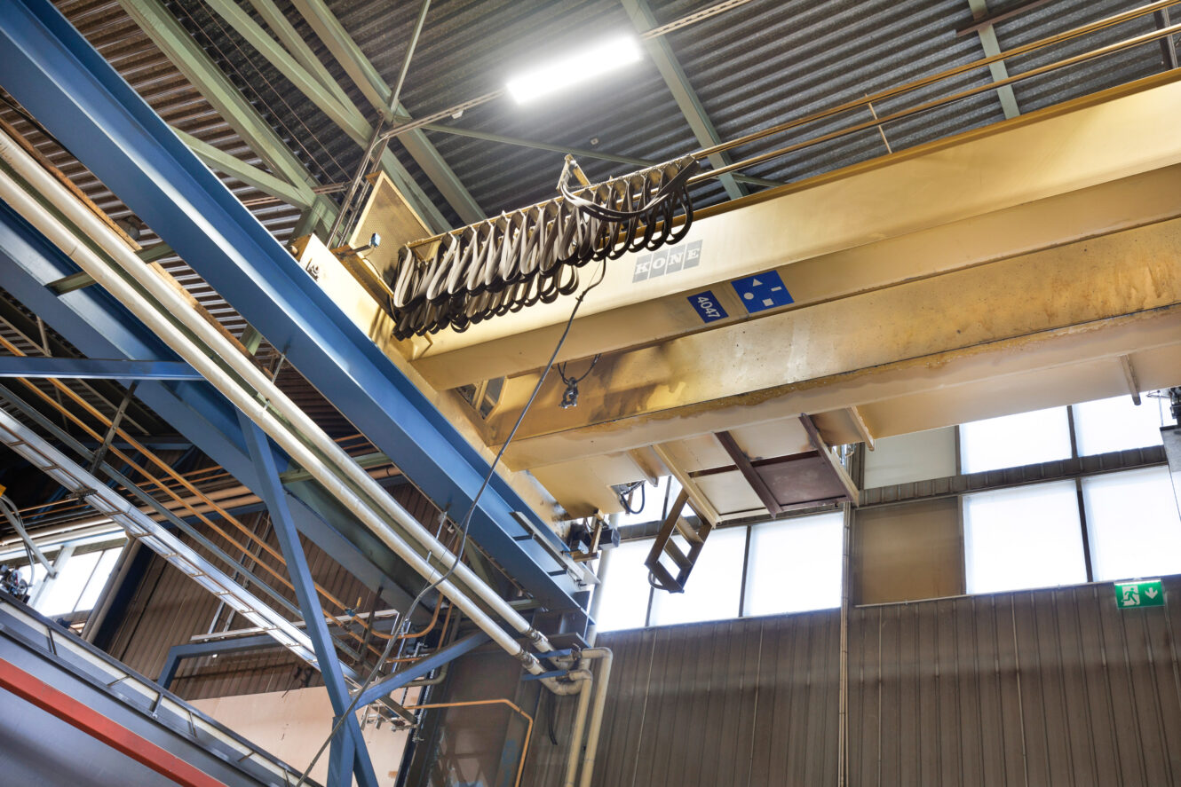 Teollinen siltanosturi koukuilla ripustettuna suuressa varastossa, taustalla näkyvät metallipalkit ja ikkunat.