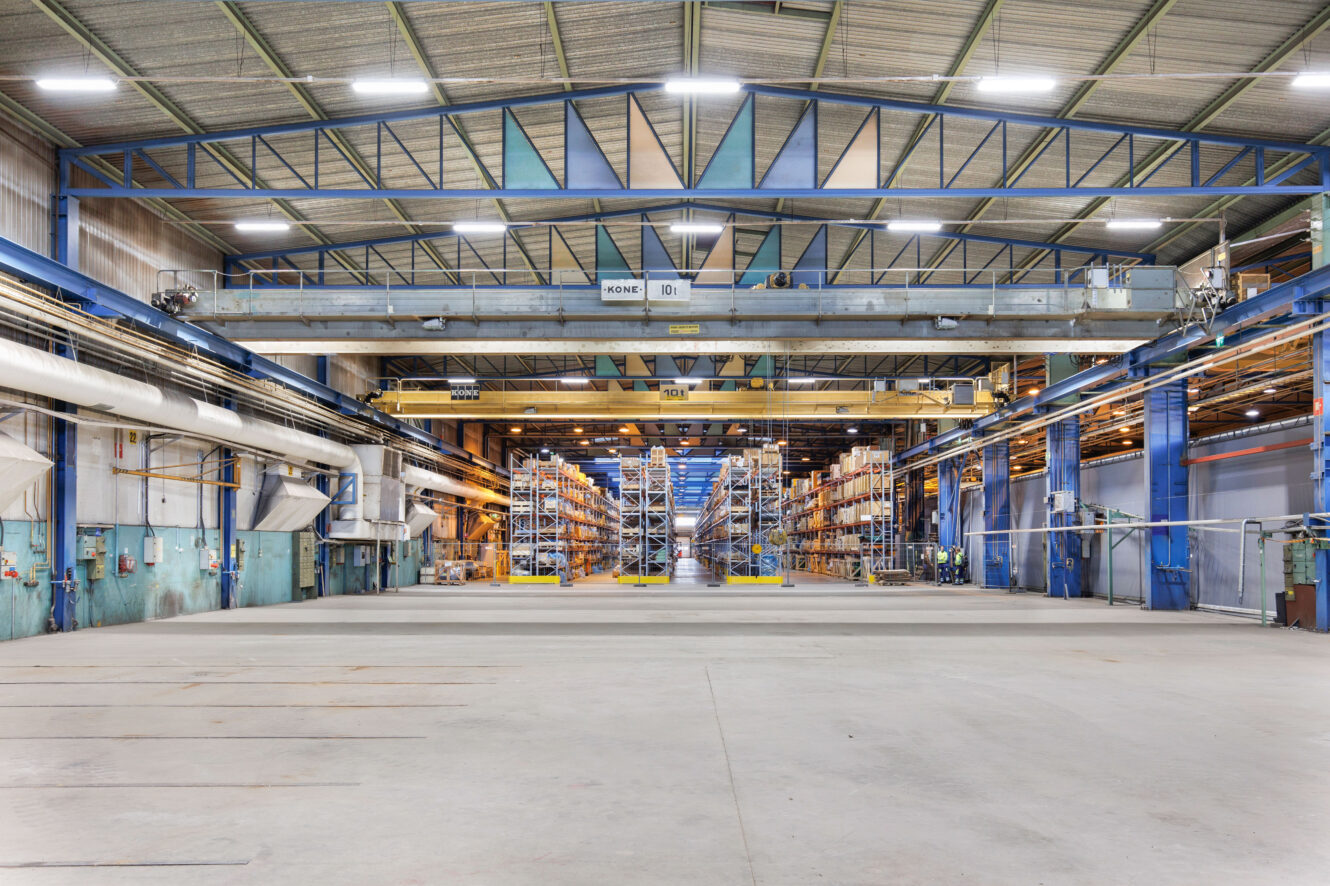 Suuren teollisuusvaraston sisätila, jossa on hyllytelineitä, nosturi ja tyhjä betonilattia, jota ympäröivät metallirakenteet ja valaisimet.