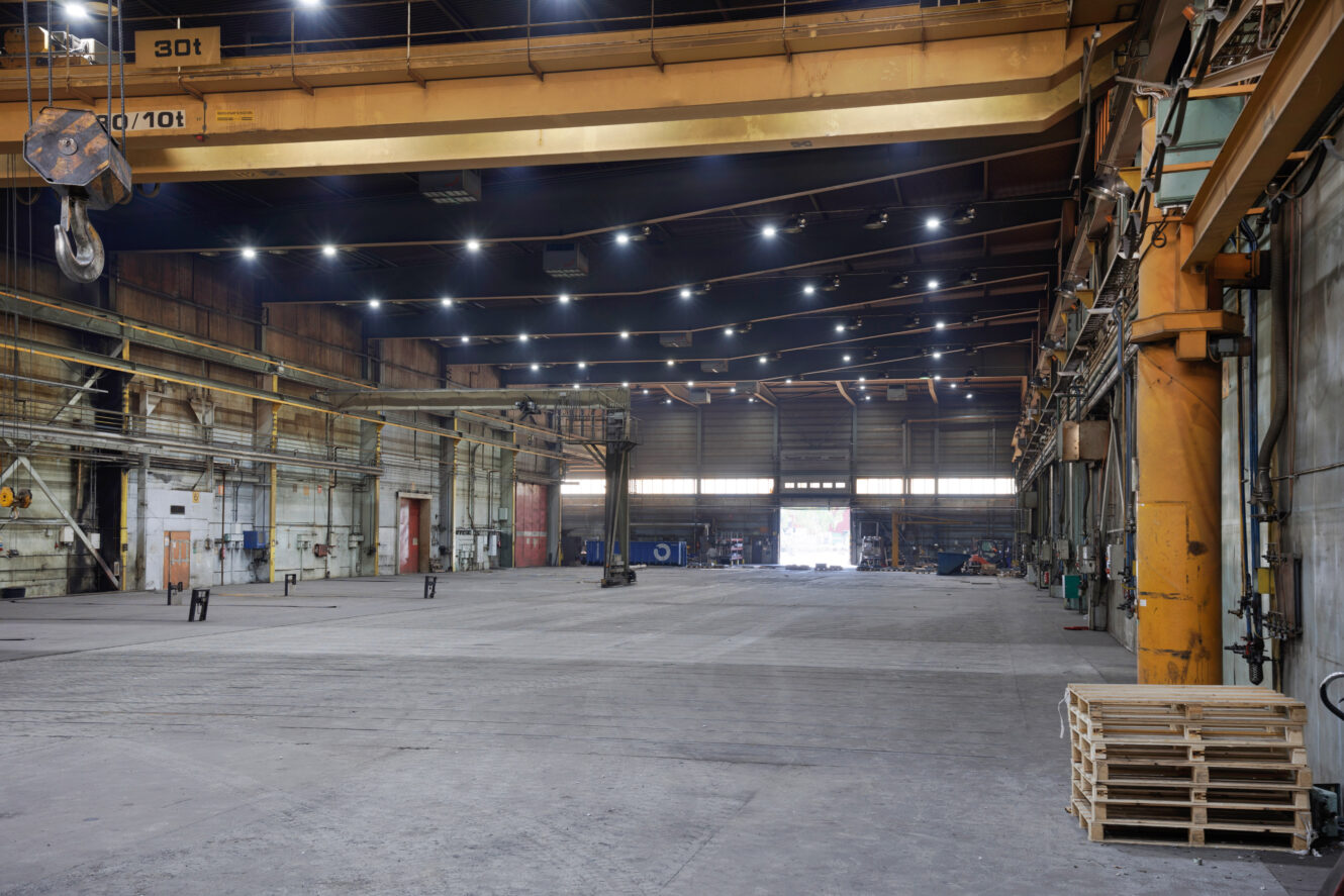 Suuri, tyhjä teollisuusvaraston sisätila, jossa on nostureita, korkeat katot, hajallaan olevia laitteita ja oikealla pinottuja puisia kuormalavoja. Kirkkaat valot valaisevat avaraa aluetta.