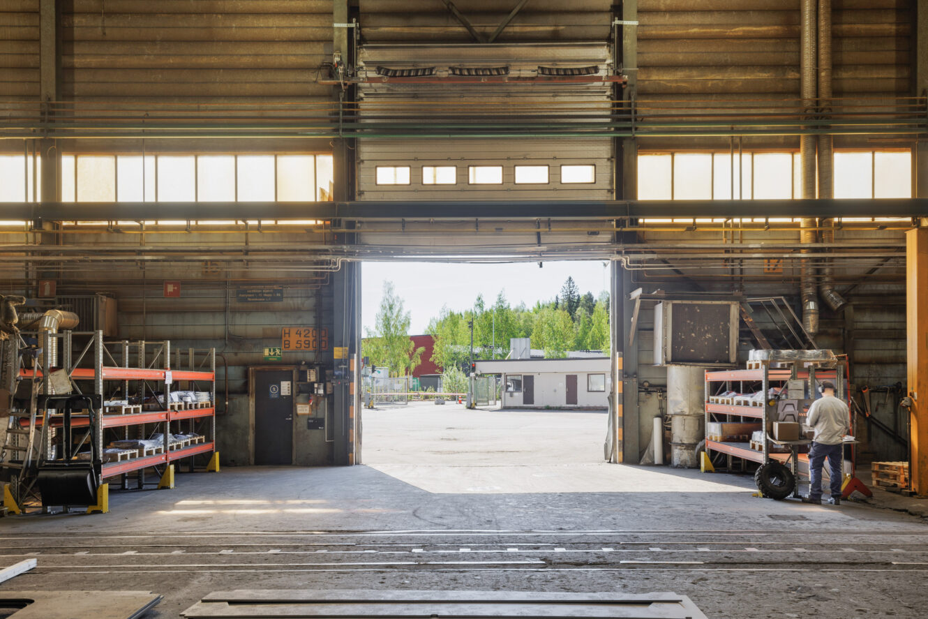 Suuren teollisuusvaraston sisätila, jossa on hyllyjä ja laitteita; henkilö seisoo sisäänkäynnin lähellä ja katsoo ulos auringonpaisteiselle pihalle, jonka ulkopuolella on rakennuksia ja puita.