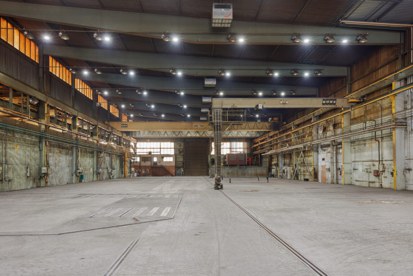 Laaja sisäkuva tyhjästä teollisuusvarastosta, jossa on betonilattiat, korkeat katot, ylävalot ja nosturijärjestelmä.
