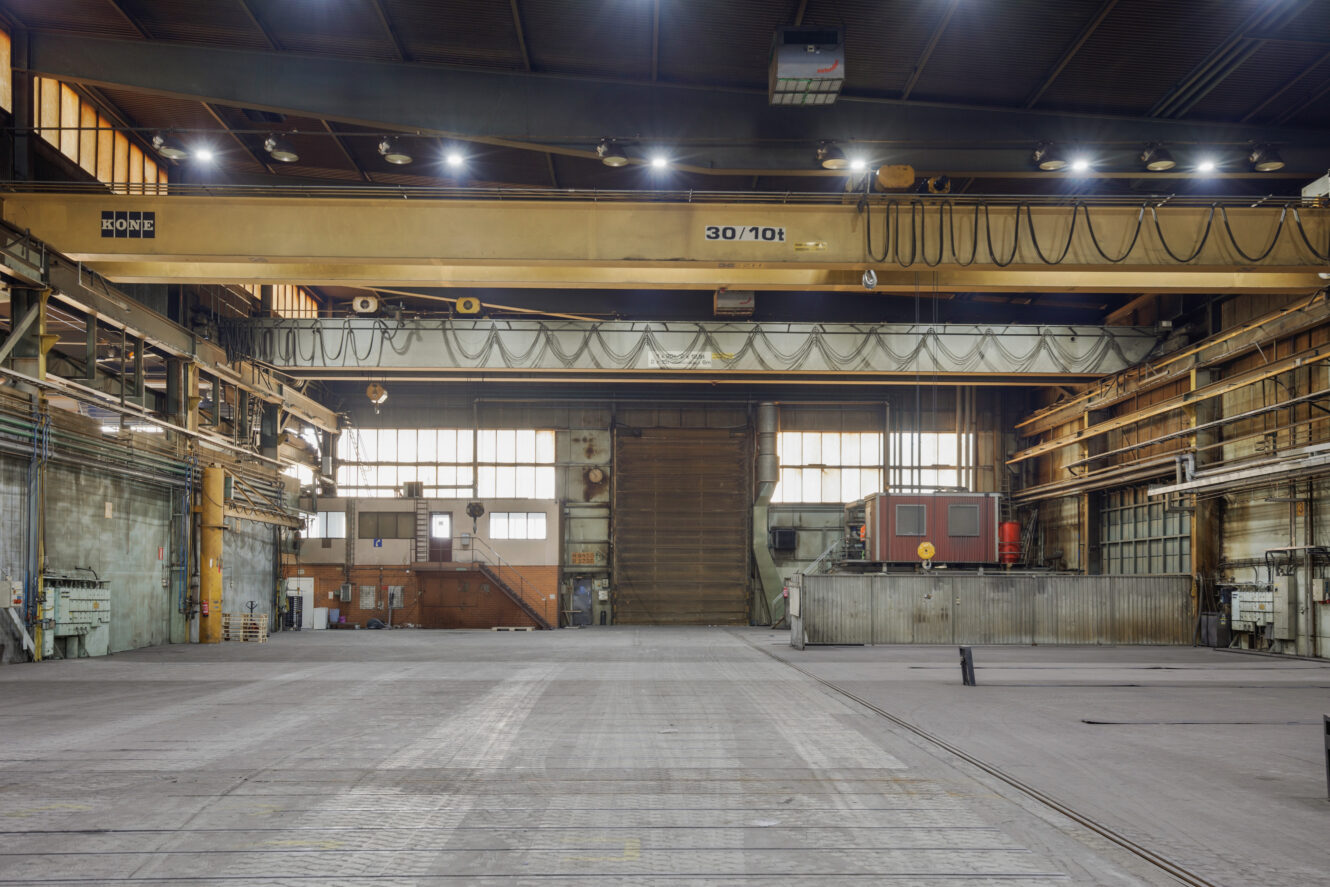 Suuren, tyhjän teollisuusvaraston sisätilat, joissa on nostureita, korkeat katot, metallipalkit ja suuret ikkunat, jotka päästävät luonnonvaloa sisään.