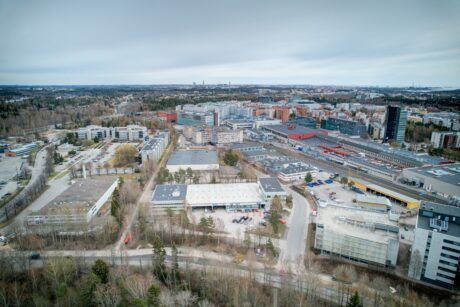 Ilmakuva kaupunkialueesta, jossa on asuin- ja liikerakennuksia, teitä ja harvaa puustoa pilvisen taivaan alla.